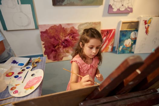 Adorable Caucasian school child, lovely little girl holding paintbrush and drawing picture on canvas, standing by a wooden easel in creative art workshop. Kids. Education. Artistic skills. Art class