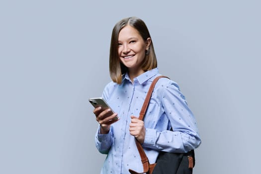 Young woman student with smartphone backpack on grey studio background. Smiling attractive female looking at camera. Using mobile applications apps for work business study leisure, technology, people