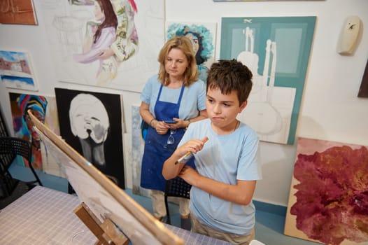 Thoughtful teenage boy artist searching for muse, inspiration and imagination while learning art, drawing on canvas in creative art studio. Female paint teacher in blue work apron on the background