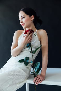 beautiful woman in a white dress sits with a red rose on a dark background