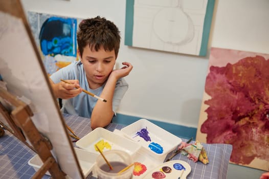 Authentic confident Caucasian teenage boy drawing picture at art class, using paintbrush and watercolor technique. Back to school. Childhood. Kid and education concept. Fine art. Elementary age school