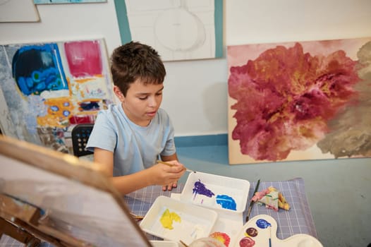 Overhead view Caucasian boy, elementary age student dipping paintbrush in water and mixing watercolors on a palette, learning art and expressing his imagination drawing picture on canvas at art class