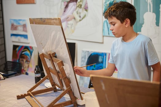 Handsome confident charming elementary age school child boy in blue casual t-shirt, standing at desk with wooden easel, holding paintbrush and painting picture with oil or acrylic paints at art class