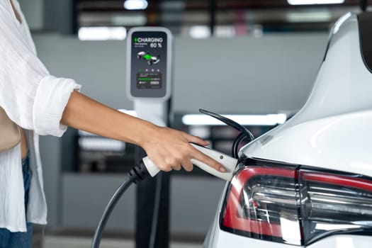 Young woman travel with EV electric car to shopping center parking lot charging in downtown city showing urban sustainability lifestyle by green clean rechargeable energy of electric vehicle innards