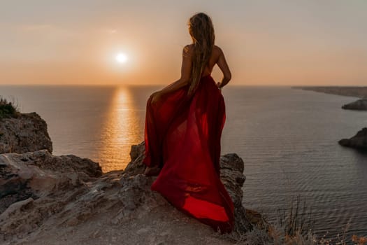 Woman sunset sea red dress, side view a happy beautiful sensual woman in a red long dress posing on a rock high above the sea on sunset