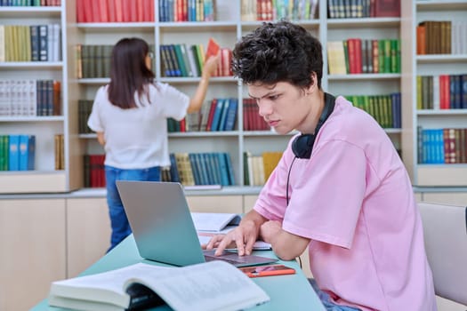 Young male student sitting with computer laptop books in college library. Guy 18-20 years old with headphones typing on laptop. Knowledge, education, youth, college university concept