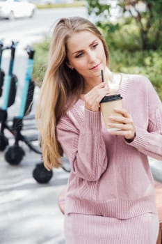 blonde woman in pink clothes drinks a drink from a mug in the park in a cafe