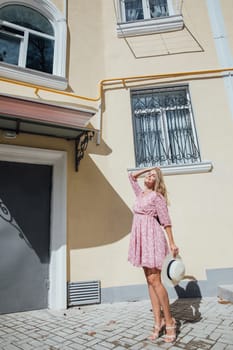 a beautiful blonde in a pink dress with a hat in her hands stands at an apartment building