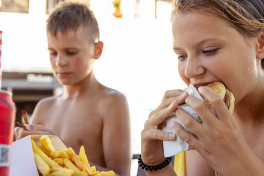 brother and sister of fair appearance eat burgers and fries on the beach, summer vacation concept. Focus on girl. High quality photo