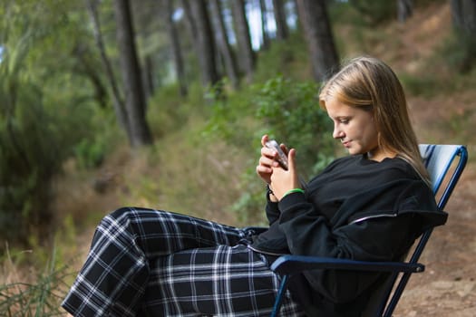 a teenage girl in casual clothes sits on a chair in a Camping and looks at the phone, a girl sits in the forest among the trees on a folding chair. High quality photo