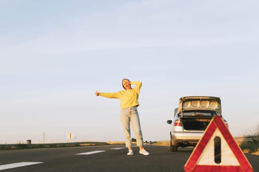 in the foreground, a red-white emergency stop sign, in the background, a girl stands with her hand raised, stops the car for help on the road, upset. Car breakdown on the road. High quality photo