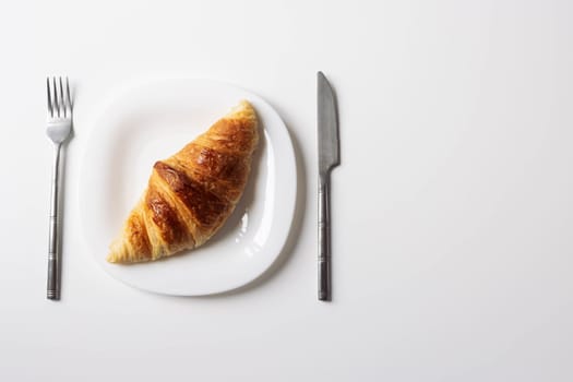 a freshly baked croissant lies on a white plate with a fork and a knife on the sides close-up isolated shot on a white... High quality photo on the right there is a place for an inscription