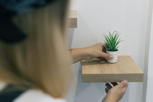 a girl with blond hair puts a green flower in a white flowerpot on a small brown shelf, close-up on a shelf, the background is blurry. High quality photo