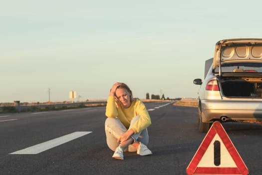 a girl in jeans of European appearance sits on the pavement near a broken car, upset, worried, there is a white-red emergency stop sign nearby. girl sits in depression. High quality photo