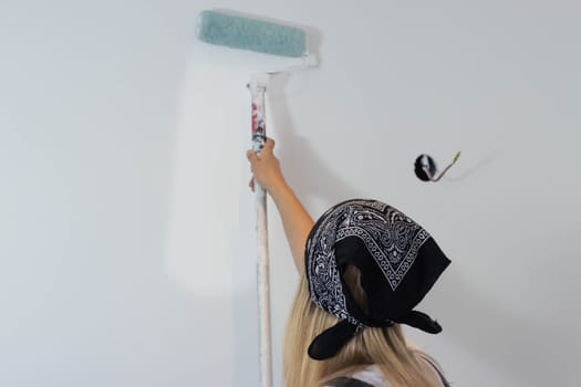 a girl with blond hair in a dark bandana, paints the wall with a roller in white at home. stands with her back to the camera. there is a place for milking the inscription. High quality photo