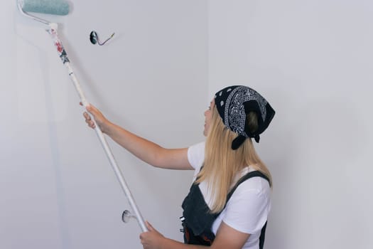 a girl with blond hair in a dark bandana, paints the wall with a roller in white at home. stands with her back to the camera. there is a place for milking the inscription. High quality photo