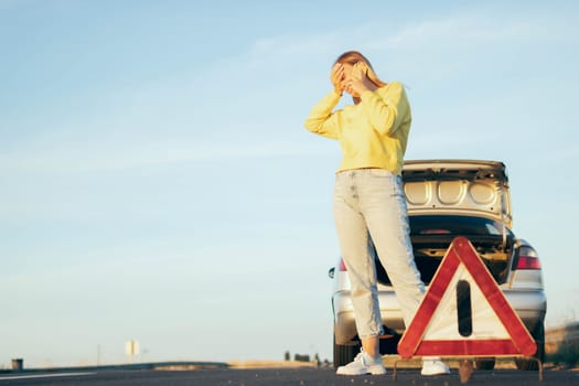 woman in a yellow sweater and jeans of European appearance stands near a broken car with an open trunk, speaks on the phone, holds her head,next to a white-red emergency stop sign. High quality photo