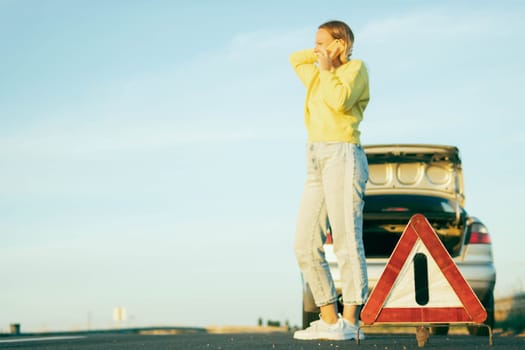 a woman in casual clothes of European appearance stands near a broken car with an open trunk, speaks on the phone, she is worried nearby there is a white-red emergency stop sign.High quality photo