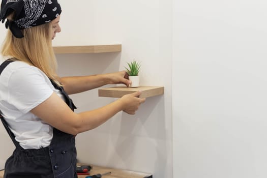 a girl with blond hair in a white T-shirt and work overalls puts a green flower in a white flowerpot on a small brown shelf, there is a place for an inscription. High quality photo