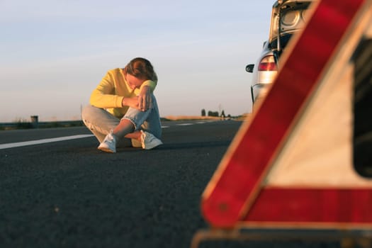 a woman in a yellow sweater and jeans of European appearance sits on the pavement near a broken car, upset, worries next to it is a white-red emergency stop sign. High quality photo