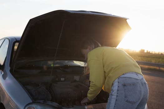 girl with blond hair in casual clothes stands with an open hood at sunset and looks at what has broken. A car breakdown on the road. There is a place for an inscription on the left. High quality photo