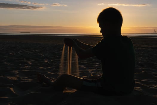 a child sits with his back to the camera on the ocean or sea at sunset plays with sand. High quality photo