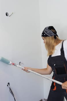 a girl with a black bandana on her head is dressed in a white T-shirt and a dark work overall, paints a white wall with a roller. Gender equality, the concept of repair. High quality photo