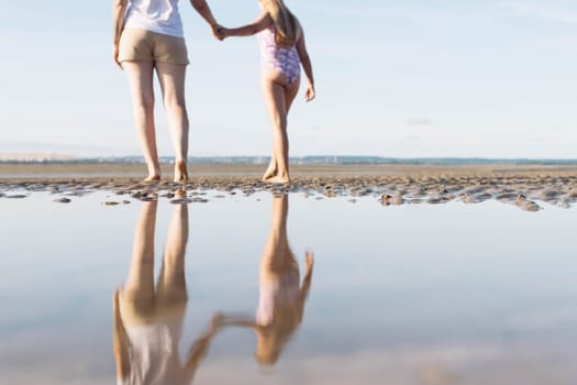 mothers in a white t-shirt and brown shorts with a daughter in a purple swimsuit walk along the beach reflection in the water in full growth photo of the legs.beach landscape. High quality photo