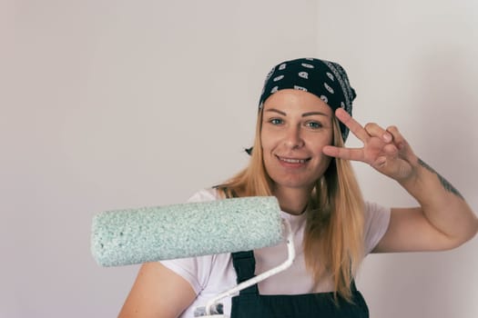 a girl of European appearance with blond hair in a bandana, paints the wall white with a roller at home. faces the camera cheerful. there is a place for an inscription. High quality photo