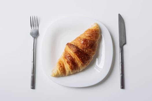 a freshly baked croissant lies on a white plate with a fork and a knife on the sides close-up isolated shot on a white... High quality photo