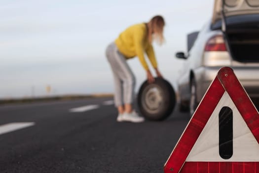 girl stands near the rear wheel of the car and rolls the spare wheel to change, the wheel has been pierced. Close-up and focus on the emergency stop sign, the background is blurred. High quality photo