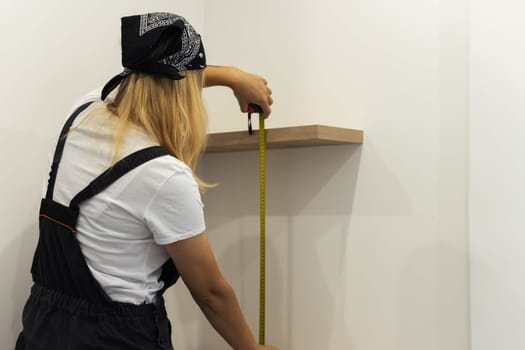 girl of European appearance in a black bandana, a white T-shirt and in a special suit in her hands a tape measure measures the shelves on the wall. The concept of equality and work. High quality photo