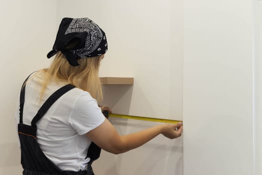 girl of European appearance in a black bandana, a white T-shirt and in a special suit in her hands a tape measure measures the shelves on the wall. The concept of equality and work. High quality photo