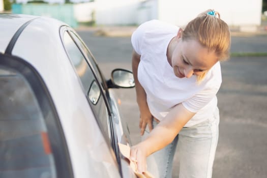 a girl of European appearance with blond hair in a white T-shirt bending over wipes the car after washing with a special yellow rag with microfiber. The concept of cleanliness. High quality photo