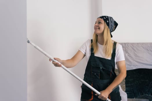 a happy girl with a black bandana on her head is dressed in a white T-shirt and a dark work overalls, paints a white wall with a roller. Gender equality, repair concept. High quality photo