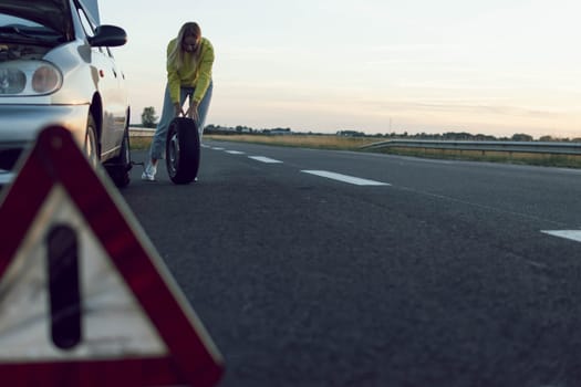 a girl in casual clothes stands near the rear wheel of the car and rolls a spare wheel to change it.the wheel in front is a temporary stop sign blurred place for an inscription. High quality photo