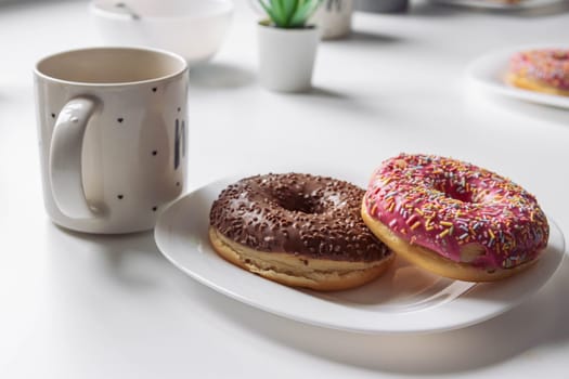 two donuts with pink and brown icing and colored sprinkles on a white plate lie on a white table, next to a white cup with black dots close-up. Photo in good quality. High quality photo