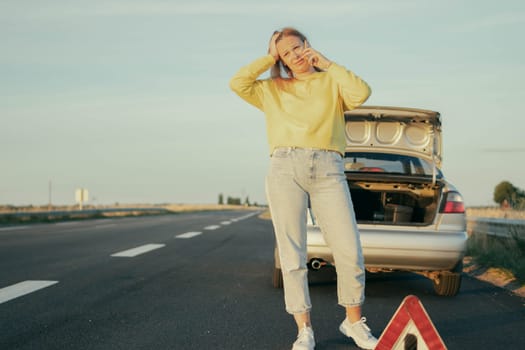 girl blond hair in a yellow sweater and jeans stands on the road near a broken car and speaks on the phone upset and holding her head,on the left there is a place for an inscription.High quality photo