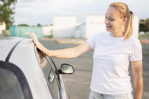 a girl of European appearance with blond hair in a white T-shirt, wipes the side windows in the car with a special yellow rag for windows. The concept of work and cleanliness. High quality photo