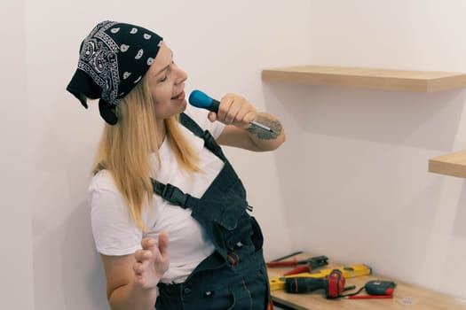 a girl in a white T-shirt and work overalls on her head with a black bandana, sings songs in a screwdriver and have fun at work on her left arm tattoo tools lie on the table. High quality photo