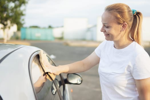 a smiling girl of European appearance with blond hair tied in a ponytail in a white T-shirt, wipes the side windows in the car with a special yellow rag while cleaning the windows. High quality photo