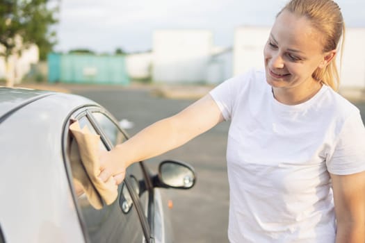 smiling girl of European appearance with blond hair tied in a ponytail in a white T-shirt, wipes the side windows in the car with a special yellow rag while milking the windows. High quality photo