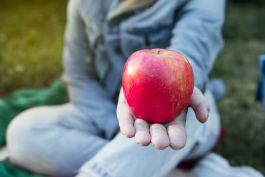 a girl sits in a denim suit in a park on the grass and holds a red beautiful apple in her hand, the focus is on the apple, the background is blurred. High quality photo