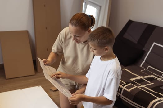 mother and son of European appearance are watching instructions on how to fold a shelf for things. They are sitting in the living room unpacking spare parts for furniture. High quality photo