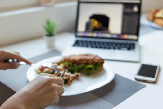 a girl sits at a white table and has breakfast, focus on hands close-up blurred background, next to it is a laptop, phone, indoor flower, there is a place for an inscription. High quality photo