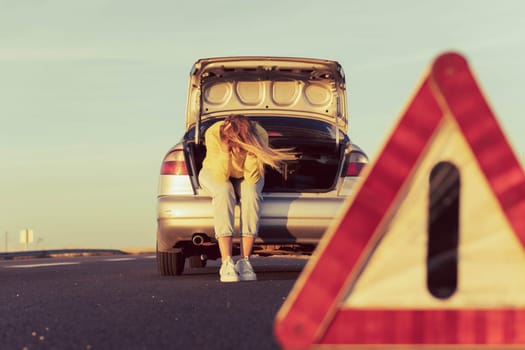 a girl with blond hair in casual clothes sits in the trunk holding her head, not knowing what to do, in the foreground an emergency stop sign close-up. High quality photo