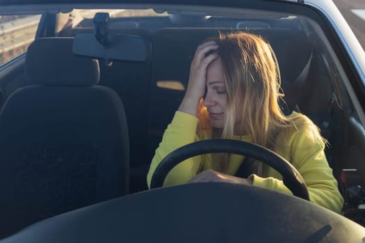 a girl of European appearance with blond hair in a yellow sweater sits behind the wheel of a car, sad, holds her head, does not know what to do, the car broke down on the road. High quality photo