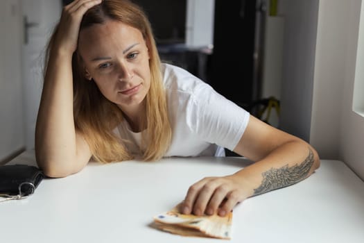 a girl of European appearance sits indoors at a white table with her head on her hands and the table is sad on the left hand of the girl with a tattoo, money lies nearby. High quality photo