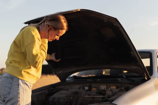 a girl with blond hair, stands with the hood of the car open and looks at what has broken. A car breakdown on the road. there is a place for an inscription on the right. High quality photo