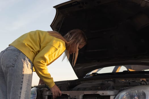 a girl with blond hair of European appearance, stands with an open hood of a car and looks at what has broken. A car breakdown on the road. there is a place for an inscription. High quality photo